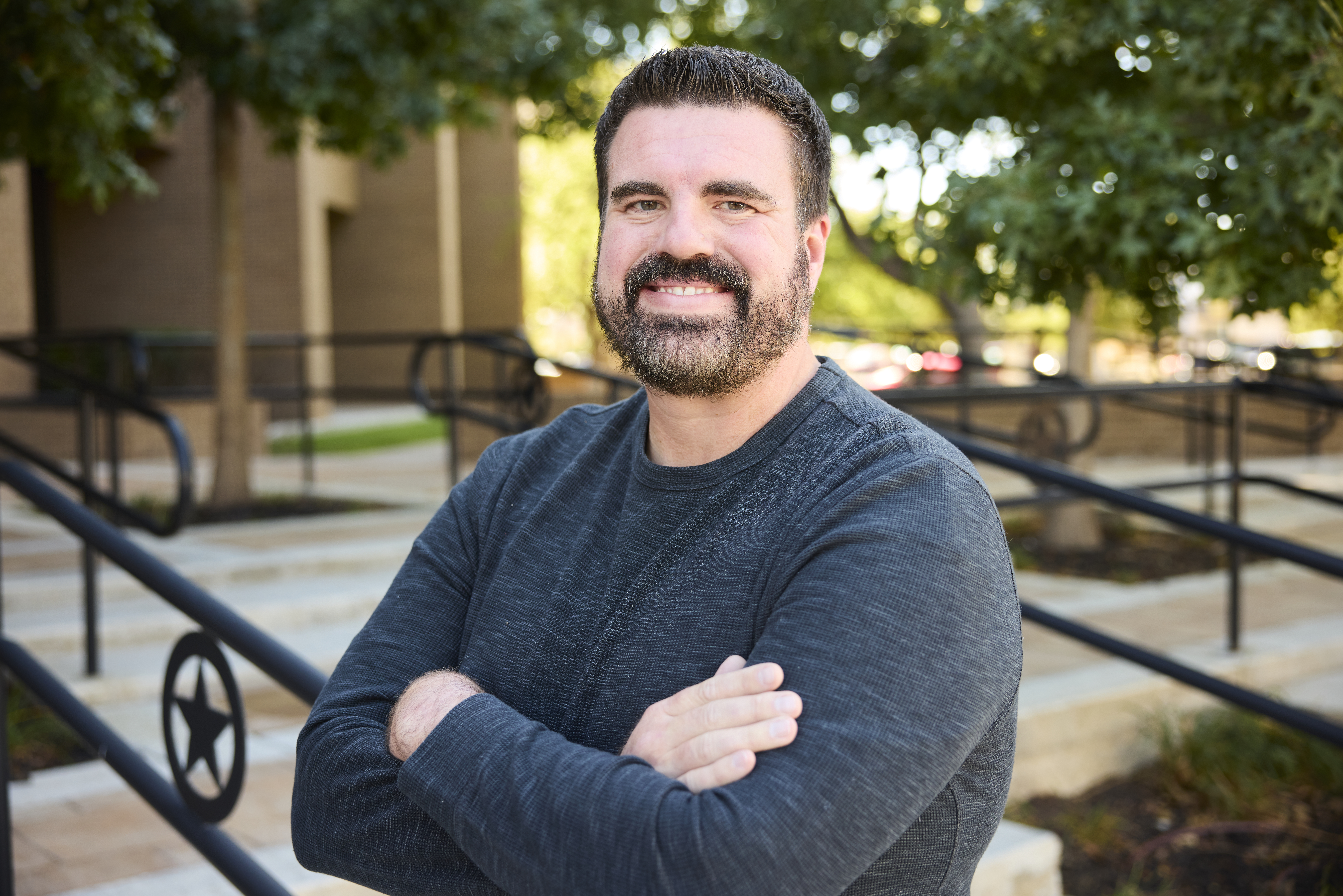 A man with dark, short-cut hair and trimmed beard is outside smiling, leaning up against the railing of steps leading up to a building. He’s wearing a dark gray shirt, arms crossed. A few trees with green leaves are in the back ground. 